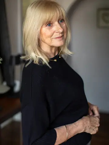 A blonde woman with blue eyes wearing a black knit mock-neck top, standing indoors with her hands clasped in front of her torso.