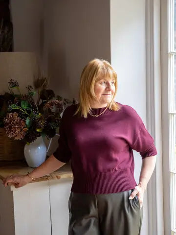 A woman with blonde hair wearing a short-sleeved burgundy knit sweater and dark olive pants, standing in a brightly lit room next to a window.