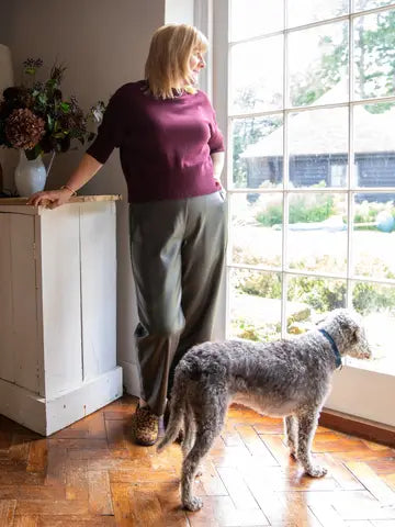 A woman wearing a burgundy short-sleeve top and olive green wide-leg trousers stands in a sunlit room next to a large window, looking out while a gray wirehaired dog stands on the parquet floor nearby.
