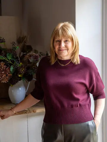 A woman with blonde hair wearing a short-sleeved burgundy knit sweater and dark gray trousers, standing indoors next to a white cabinet with a vase of dried flowers.