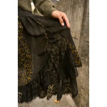 A close-up of a black skirt with layered ruffles and gold floral lace trim, worn by a person whose hand is resting on the fabric, set against a rustic concrete wall.