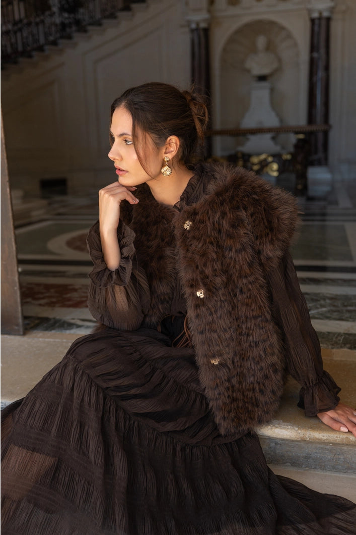 Woman in a dark fur coat and dress sitting on steps in an elegant interior setting.