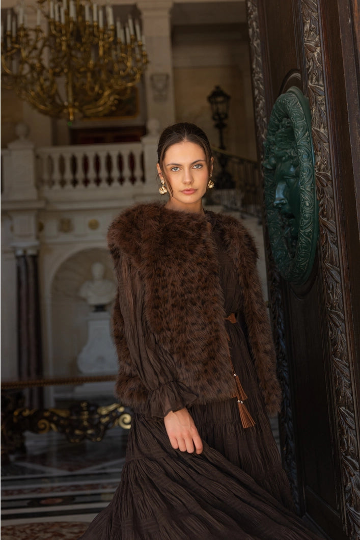 Woman in a brown fur coat standing in an ornate room with a chandelier.