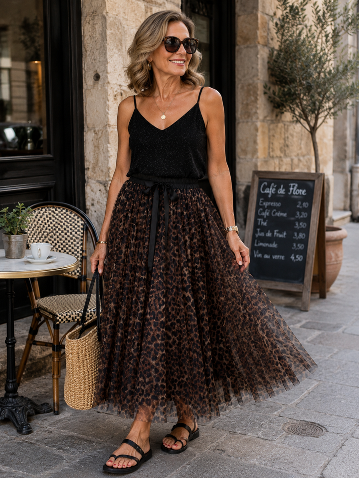 A woman stands on a stone sidewalk in front of a cafe, wearing a black sleeveless top and a long brown leopard-print tulle midi skirt with a black tie belt. She is holding a woven straw tote bag.