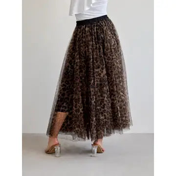 A leopard-print tulle midi skirt with a black waistband, shown on a model from the waist down wearing clear high-heeled sandals against a plain light gray studio background.