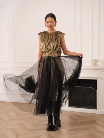 Woman wearing a gold sequin top and black tulle skirt in a room with white walls and a fireplace.