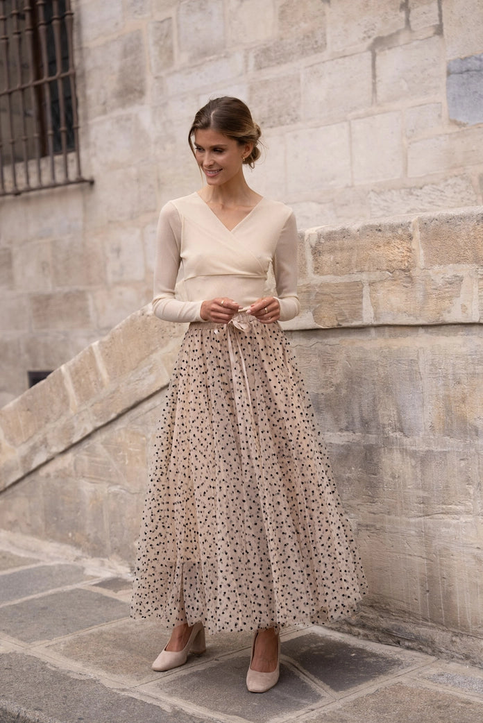 Woman in a long, light-colored dress with a patterned skirt standing against a stone wall.
