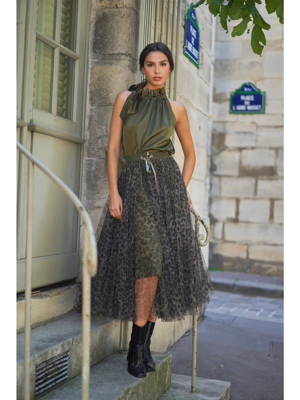 Woman in a green top and patterned skirt standing on a street in an urban setting.