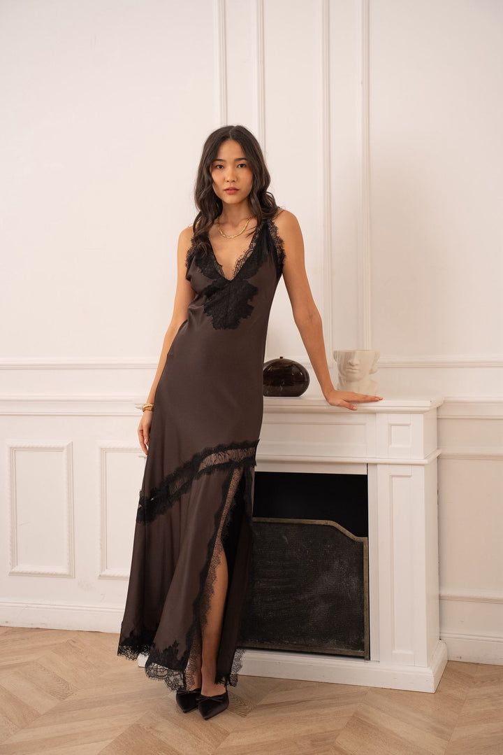 Woman in a long, dark brown dress standing in a room with white paneled walls and a fireplace.