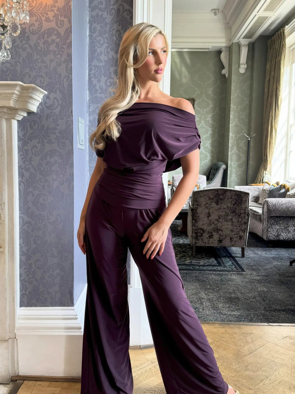 Woman wearing a purple off-shoulder jumpsuit in an elegant room.