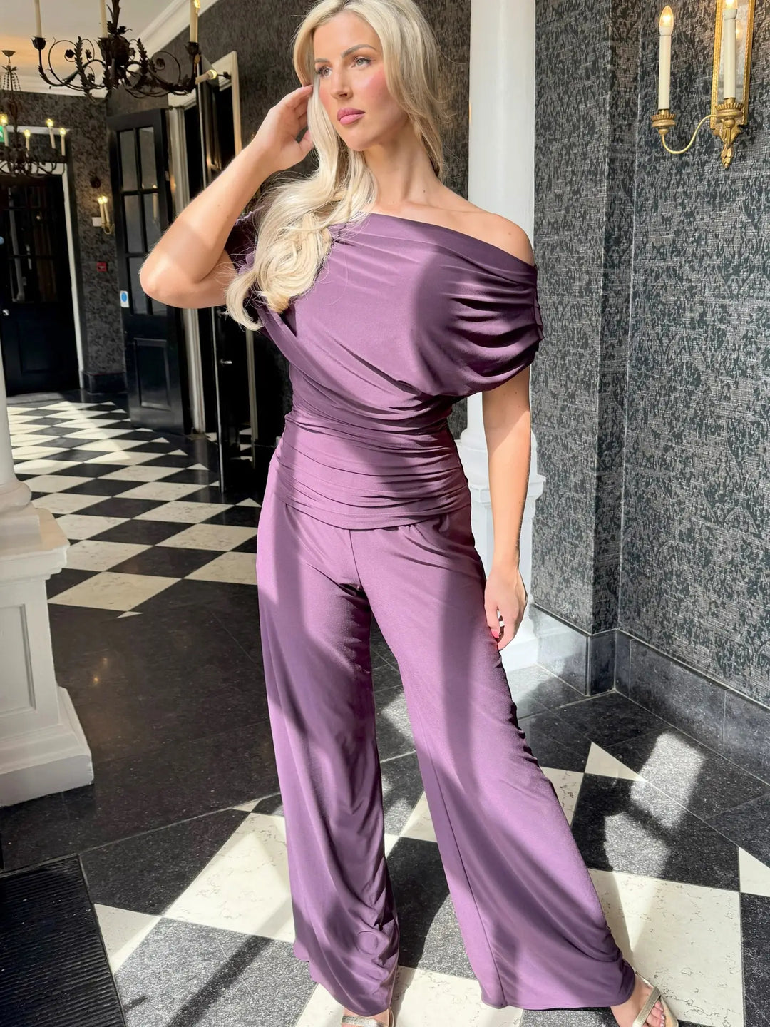Woman in a purple jumpsuit standing in an elegant interior setting with checkered floor and marble walls.
