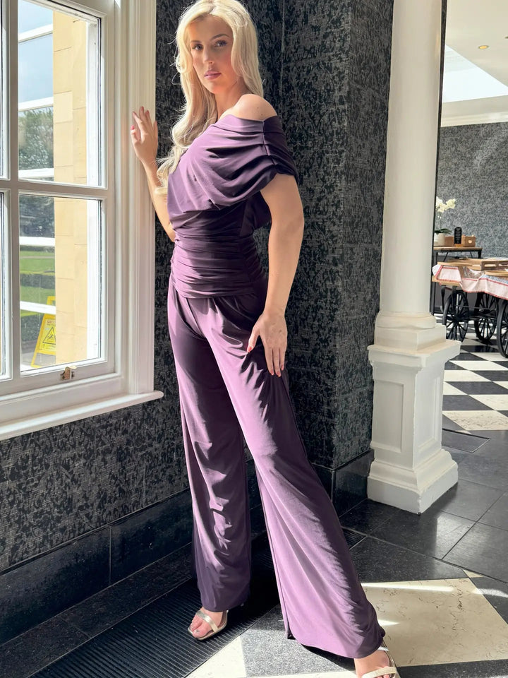 Woman in a purple off-shoulder top and pants standing indoors near a window.