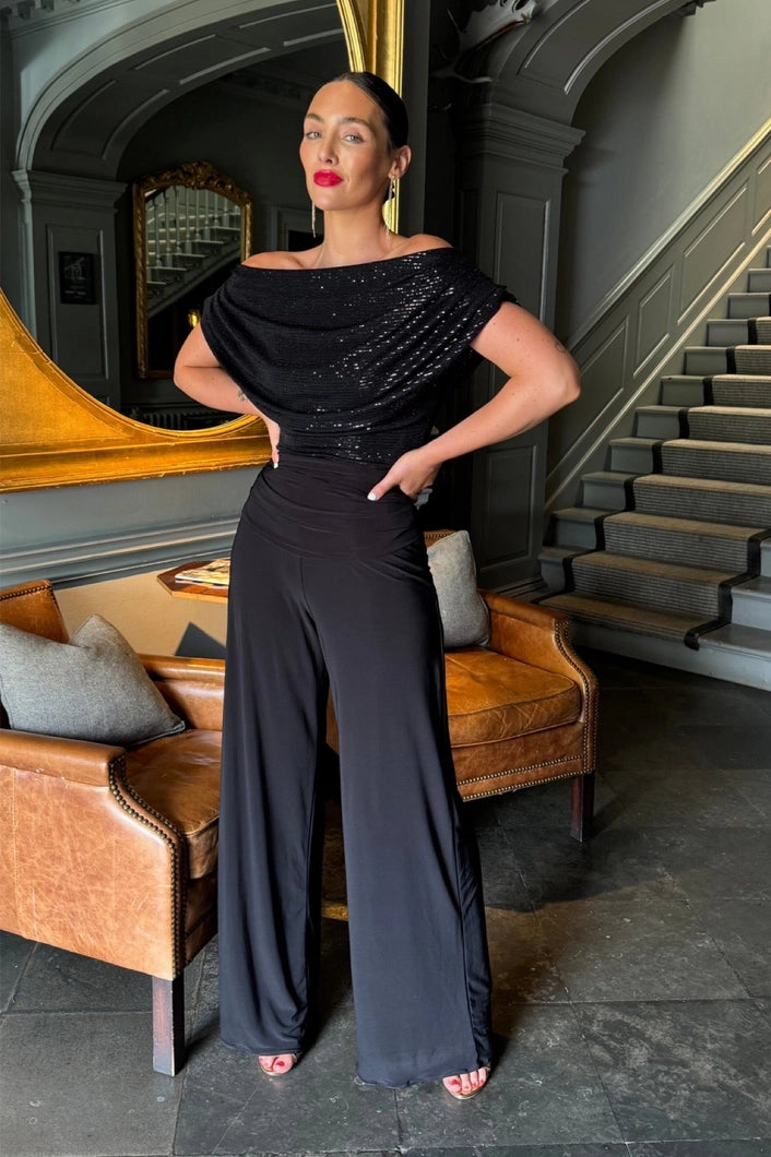Woman in a black outfit standing in an elegant interior setting with a staircase and mirror.