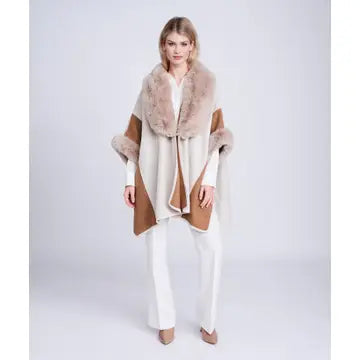 A model wearing a beige and brown wrap poncho with a plush faux-fur collar, styled with white pants and beige heels against a light gray studio background.