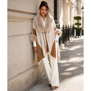 A woman stands on a city sidewalk leaning against a beige stone building, wearing a long cream and tan wrap-style coat with a large fluffy light beige fur collar and cuffs, paired with white wide-leg pants.