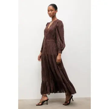 A woman modeling a long-sleeved, dark brown lace maxi dress with a tiered skirt, paired with dark brown heeled sandals, against a plain off-white studio background.