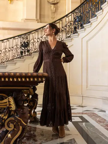 A woman wearing a dark brown lace long-sleeve maxi dress stands on a marble staircase landing next to an ornate wooden table in a grand interior.