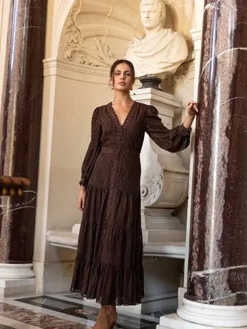 A woman models a dark brown long-sleeve midi dress with a textured crinkle fabric, standing in an elegant interior with marble columns and a classical bust.