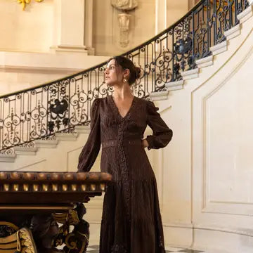 A woman wearing a dark brown long-sleeved lace dress stands in front of an ornate indoor staircase with a black iron railing.