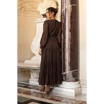 A woman wearing a dark brown long-sleeve midi dress with a pleated skirt, standing in an elegant interior next to a marble column.
