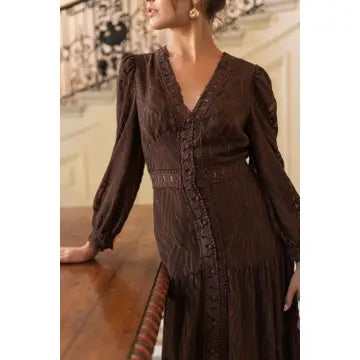 A woman wearing a dark brown long-sleeve midi dress with a V-neckline, textured pattern, and decorative button details down the front, standing indoors next to a wooden table.