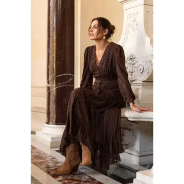 A woman wearing a dark brown lace long-sleeve maxi dress and brown leather cowboy boots sits on a white stone bench in an elegant indoor setting with columns.