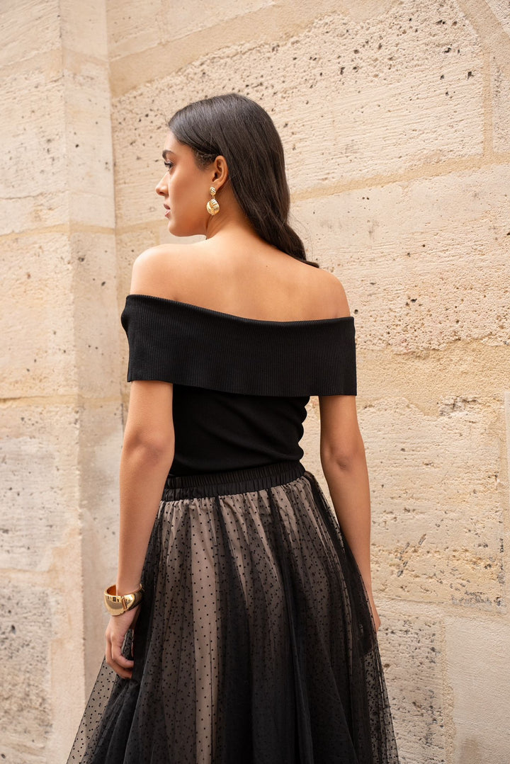 Woman wearing a black off-shoulder top and patterned skirt against a stone wall.
