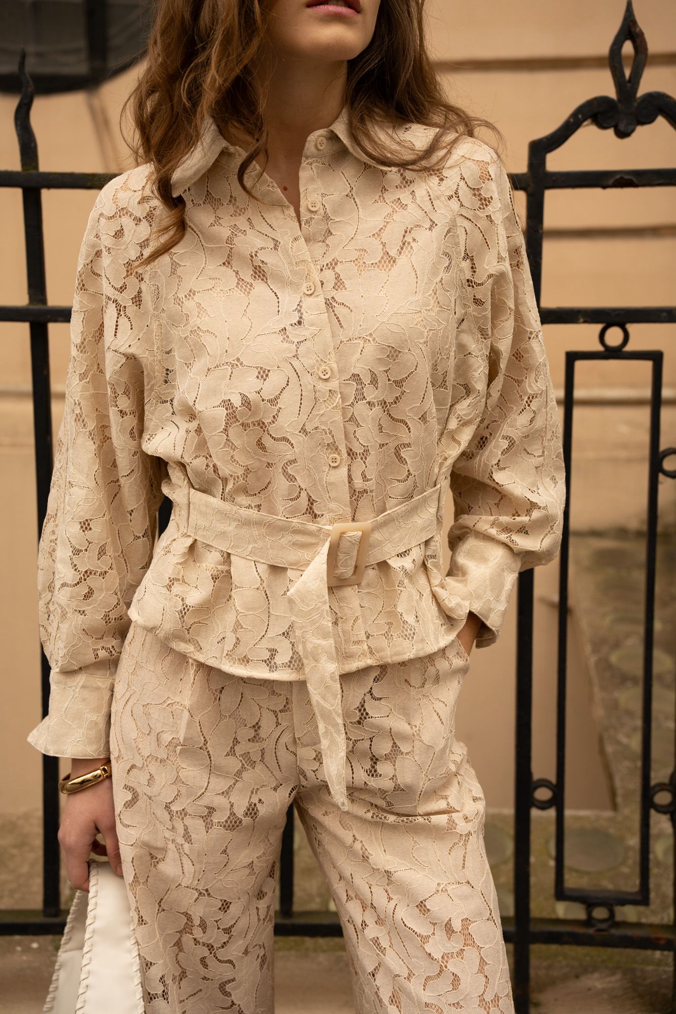 Person wearing a beige lace outfit in front of a black metal gate.