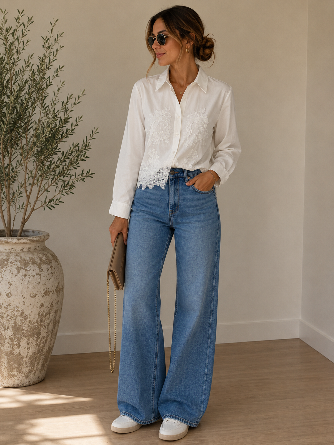 A woman wearing a white long-sleeve blouse with lace trim and high-waisted blue wide-leg jeans, standing in a bright indoor setting with a large vase and plant in the background.