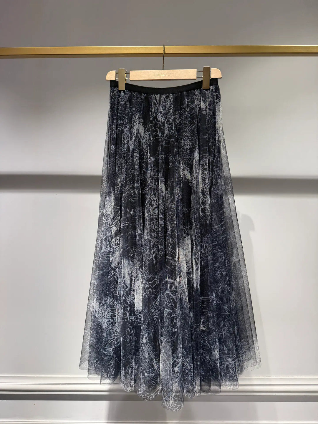A pleated midi skirt in a black and white marble or tie-dye pattern, hanging from a wooden hanger on a gold-colored clothing rack against a white wall.