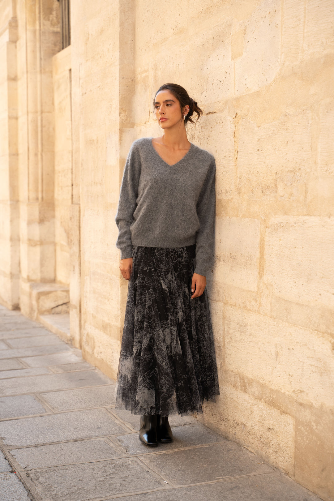 A woman stands against a beige stone wall wearing a gray knit V-neck sweater and a long black tulle skirt with a gray marbled pattern, paired with black boots.