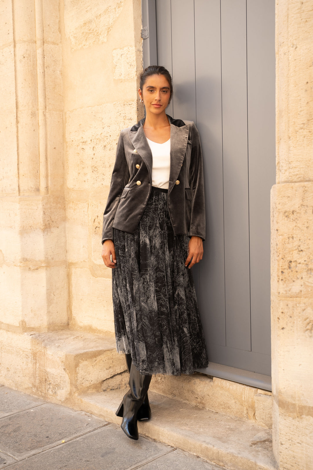 A woman stands outdoors against a beige stone wall and gray door, wearing a dark gray velvet blazer with gold buttons over a white top, a dark gray patterned midi skirt, and black ankle boots.
