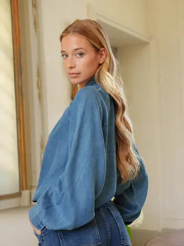 A woman with long blonde hair wearing a blue suede long-sleeve top and blue jeans, photographed indoors against a light cream-colored wall.