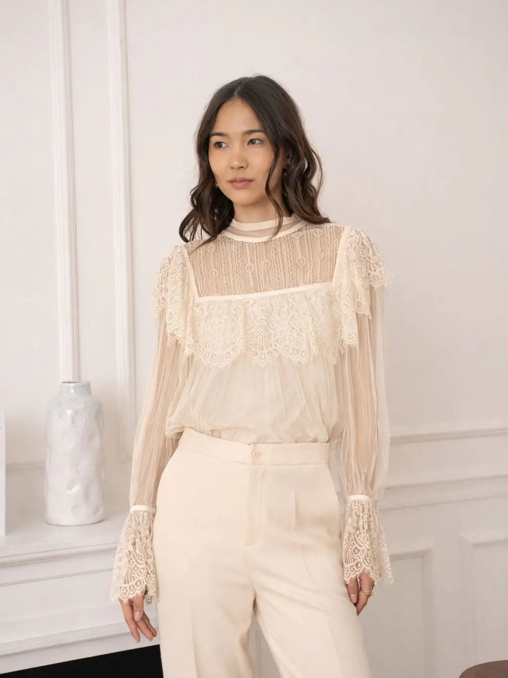 Woman wearing a beige lace blouse and pants against a white wall.