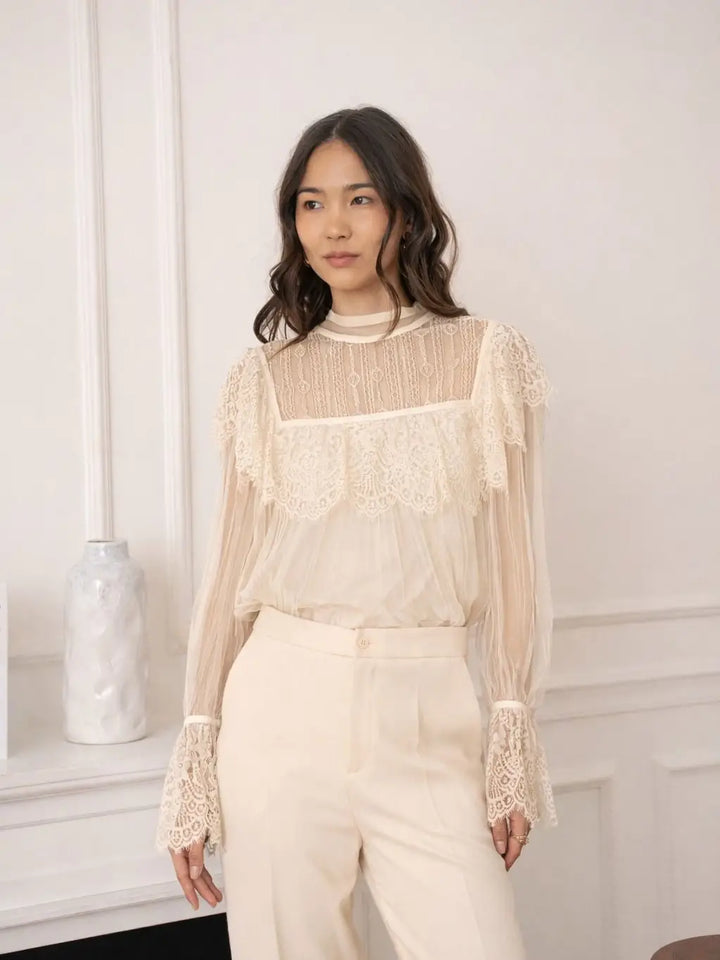 Woman wearing a beige lace blouse and pants against a white wall.
