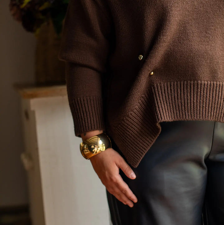 A close-up lifestyle shot of a person wearing a brown knit cardigan with gold buttons, paired with black leather pants and a chunky gold cuff bracelet.