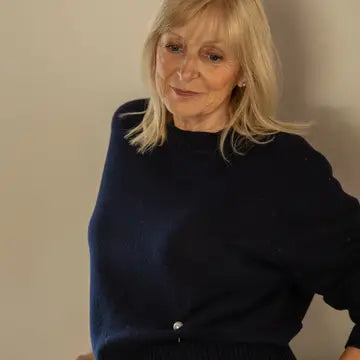 A woman with blonde hair wearing a dark navy blue knit sweater, photographed against a plain beige wall.