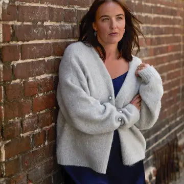 A woman wearing a fuzzy light gray cardigan with silver buttons over a navy blue top, leaning against a red-brown brick wall.
