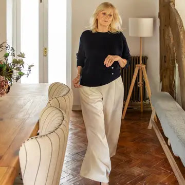 A woman walking indoors wearing a dark navy long-sleeve top and light beige wide-leg trousers in a rustic room with wood floors and furniture.