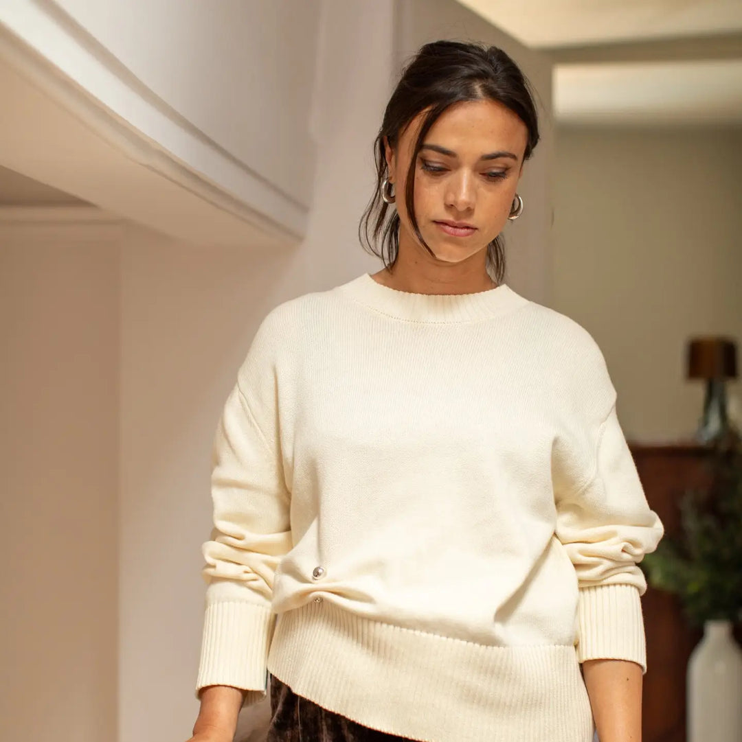 A woman wearing a cream-colored knit sweater with ruched sleeves and a ribbed hem, standing in a softly lit indoor setting.