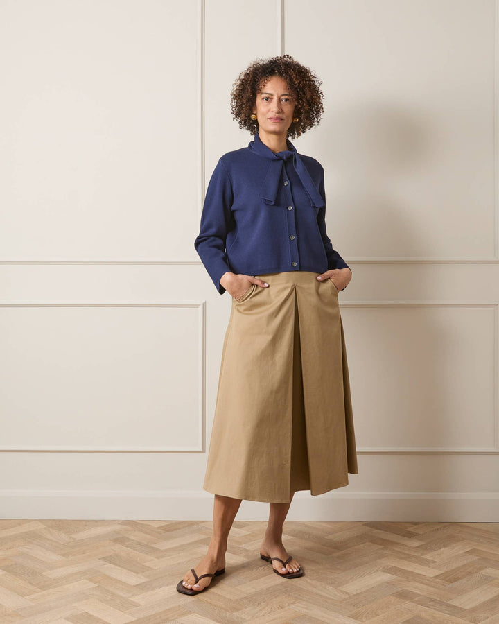 Woman wearing a blue shirt and beige skirt standing against a white paneled wall.