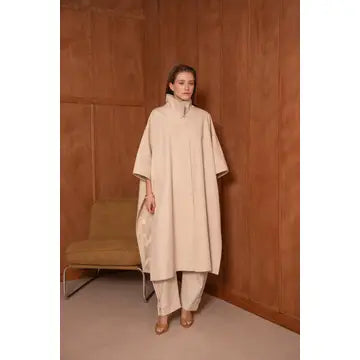 Woman wearing a beige poncho standing against a brown wall.