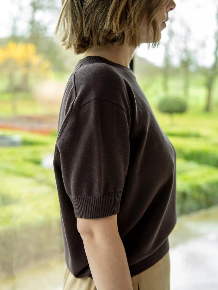 Person wearing a brown sweater standing in front of a window with a garden view