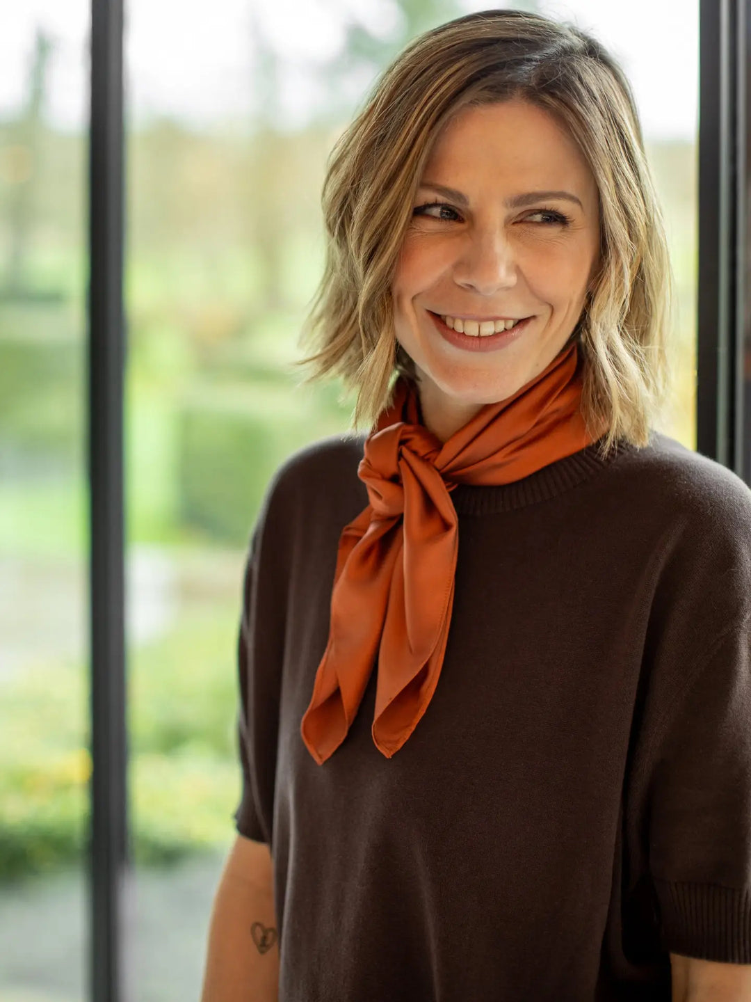 Woman wearing an orange scarf and brown shirt with a blurred outdoor background