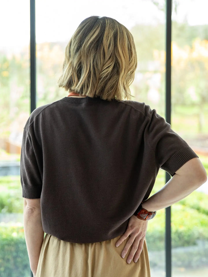 Person wearing a brown sweater and beige pants standing in front of a glass door with a blurred outdoor background.