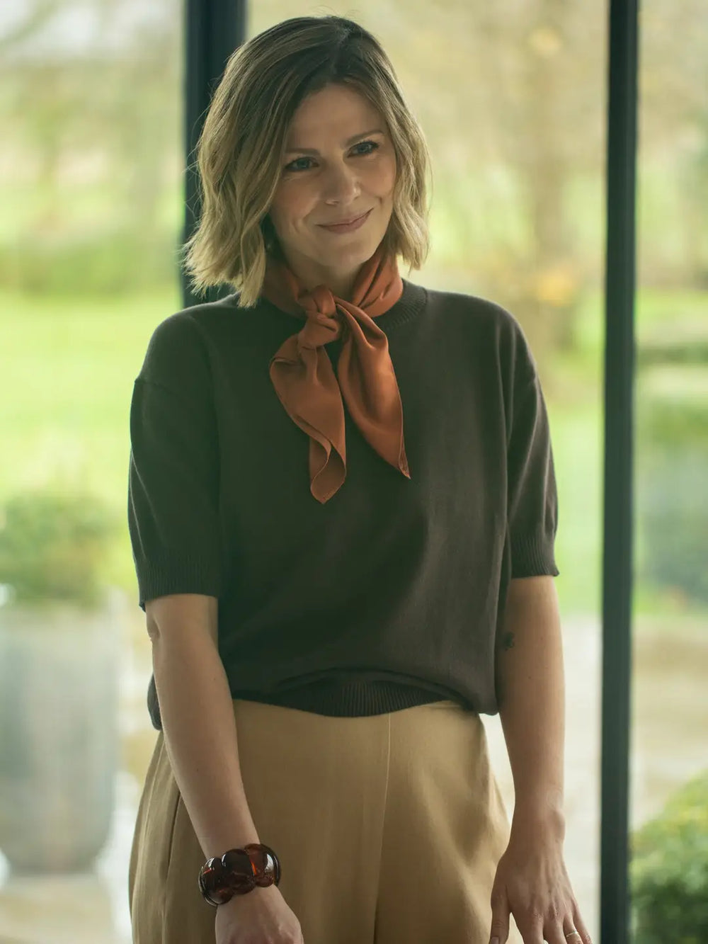 Woman wearing a green top and beige pants with an orange scarf, standing indoors.
