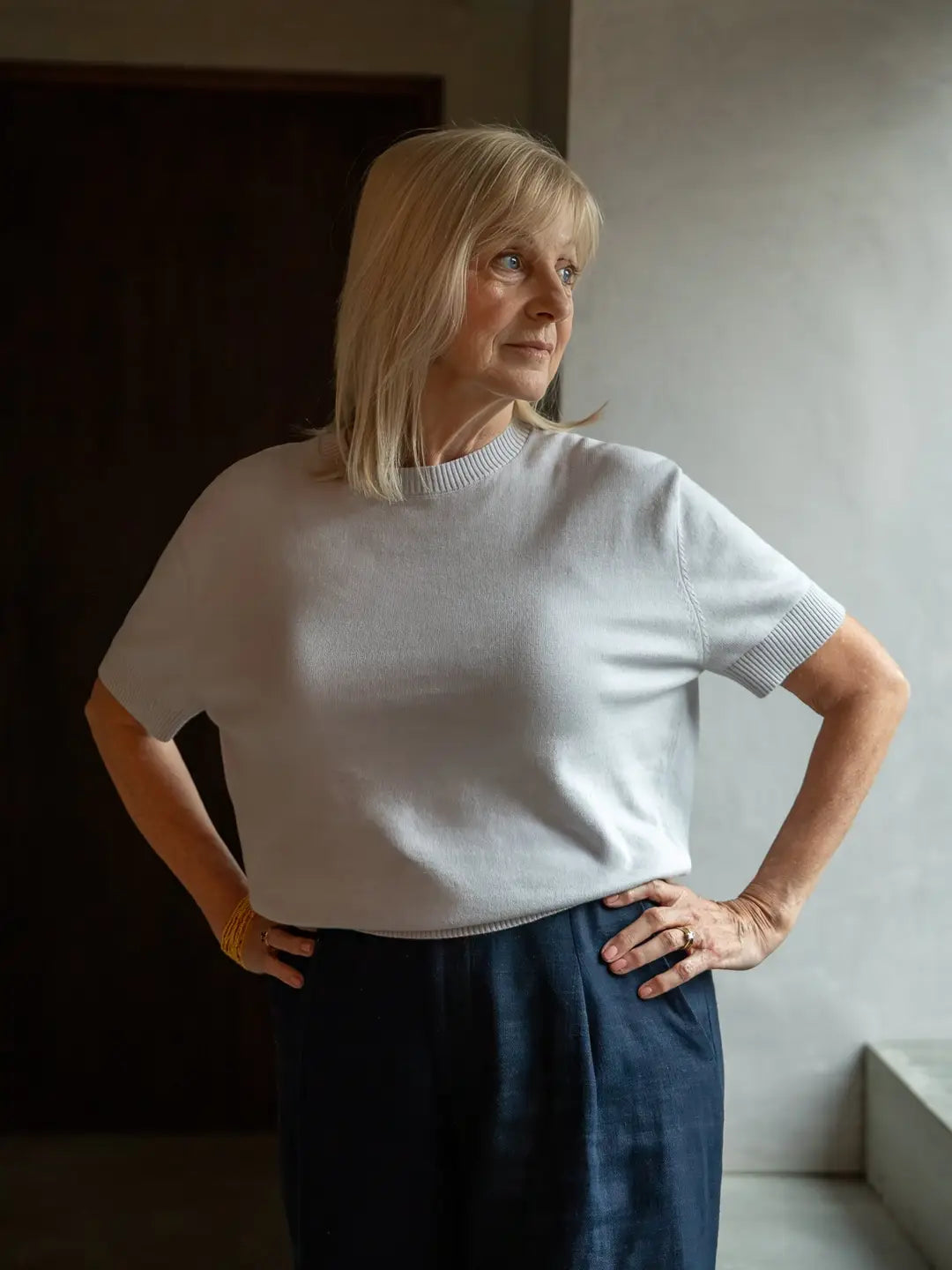 Woman wearing a white sweater and dark pants standing against a neutral wall.