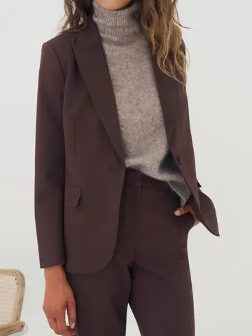 A woman wearing a dark brown tailored blazer with matching trousers, layered over a light gray turtleneck sweater, shown against a plain light background.
