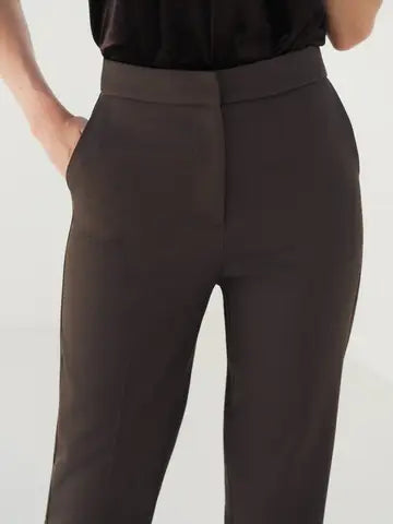 A close-up of a model wearing dark brown tailored trousers with a wide waistband and side pockets, paired with a black top against a light gray background.