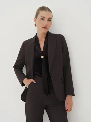 A woman modeling a tailored black blazer over a black satin blouse and black trousers, standing against a plain light gray wall.